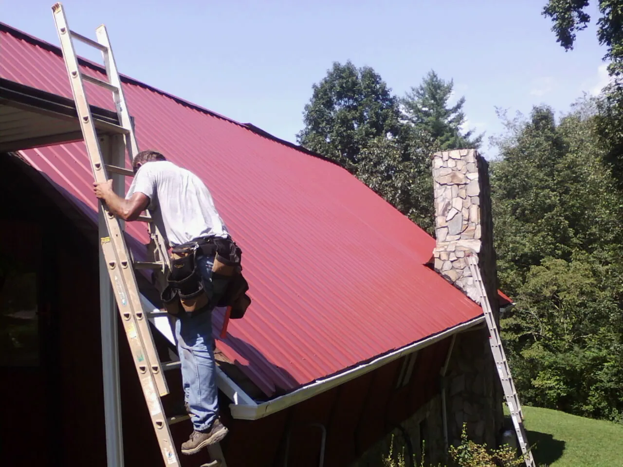 Roofer on a ladder beside a red metal roof and stone chimney — Soffit & Fascia Repair by Roofing Brothers in Fountain Valley
